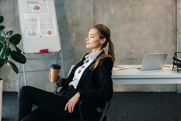 Young businesswoman seating on chair with closed eyes and holding paper cup of coffee - Foto, immagini