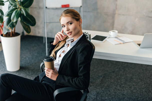 young businesswoman sitting on chair with paper cup of coffee and touching face  - Foto, Imagen