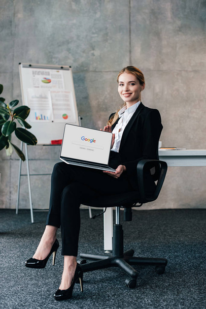 pretty smiling businesswoman sitting in chair and holding laptop with google on display - Fotoğraf, Görsel
