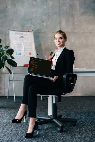 Lächelnde Geschäftsfrau sitzt auf Stuhl und hält Laptop mit Programmiercode auf dem Bildschirm - Foto, Bild