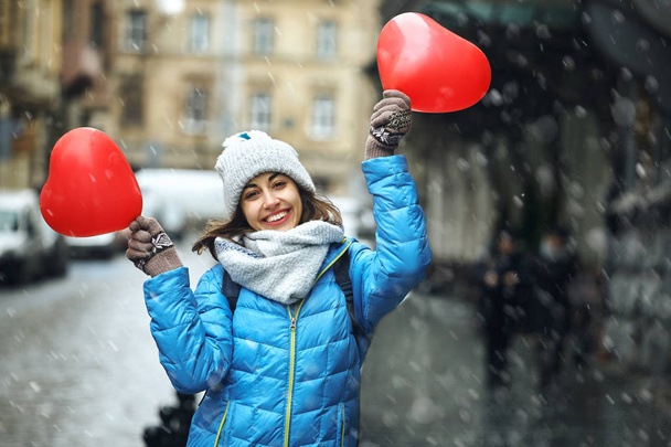 happy young woman in warm winter clothes, knitted cap and scarf is standing outdoors with a heart shaped red balloons - Photo, image