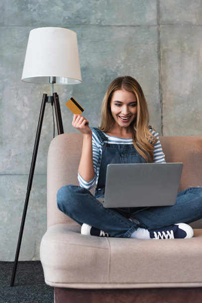 szczęśliwy, piękna kobieta, trzymając w ręku karty kredytowej i siedzi na kanapie z laptopem  - Zdjęcie, obraz