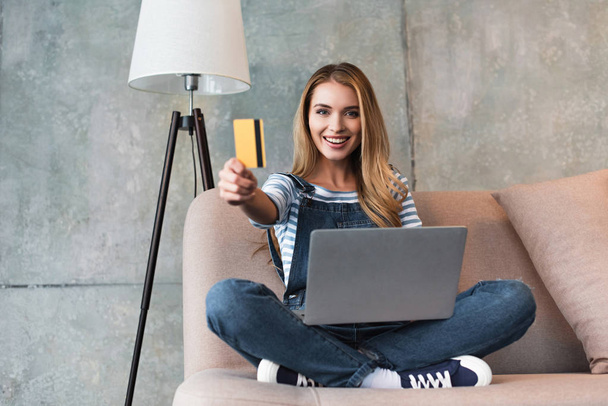 beautiful woman showing credit card and sitting on sofa with laptop  - Photo, Image
