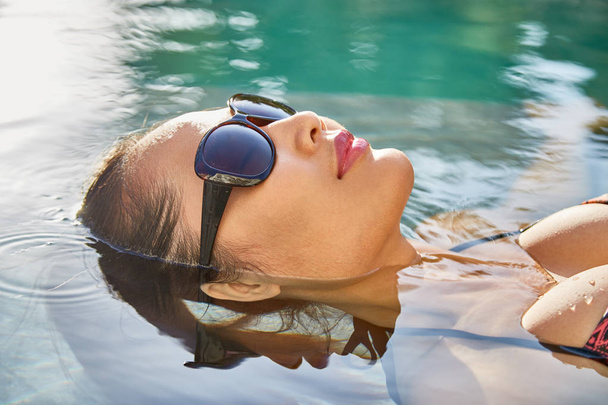 Mujer joven en gafas de sol flotando sobre su espalda en la piscina
 - Foto, imagen