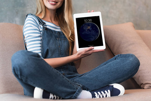 cropped view of girl sitting on sofa and showing screen with earth of digital tablet - Foto, Imagem