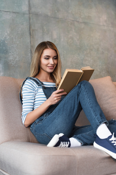 young smiling girl sitting on pink sofa and reading book - Fotoğraf, Görsel