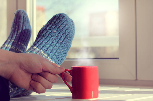 feet in warm socks and a mug with a hot drink on the table in front of window / warming atmosphere of home holiday - Fotografie, Obrázek