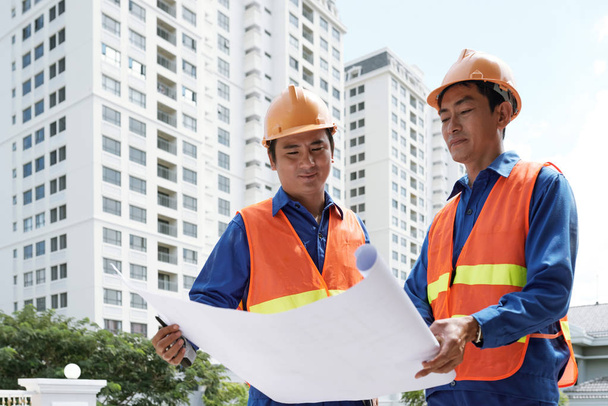 Hymyilevät aasialaiset rakennustyöntekijät tapaavat rakennuksen ulkopuolella ja keskustelevat suunnitelmasta
 - Valokuva, kuva