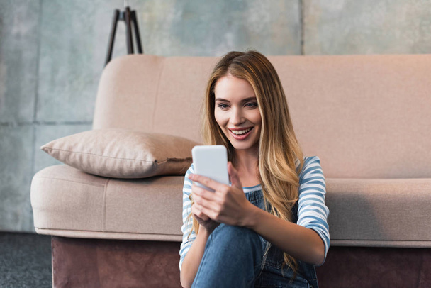 young woman smiling, using smartphone and sitting near pink sofa - Foto, immagini