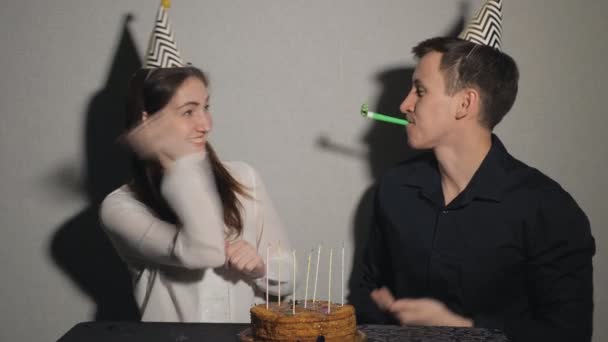 Pretty girl and man celebrates a holiday, sits at a table with a cake and a candles playing with party blowers - Footage, Video
