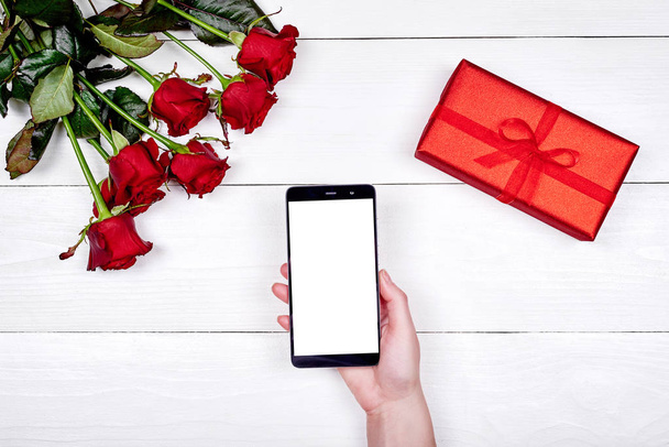 Mobile smart phone with blank screen in woman hands, bouquet of roses and gift box on white background, copy space. Valentines day, Womens Day, Mothers Day. Top view, flat lay  - Foto, immagini