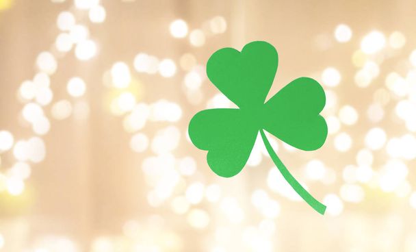 fortune, luck and st patricks day concept - green paper shamrock over festive lights on beige background - Photo, image
