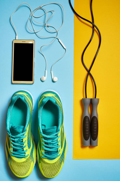 Pair of sport shoes, skipping rope and cellphone with earphones. New sneakers on blue and yellow pastel background, copy space. Overhead shot of running foot wear. Top view, flat lay - Photo, Image