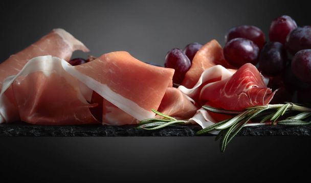 Prosciutto with grapes and rosemary on a black background. Closeup, copy space. - Photo, Image