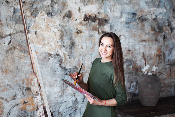 Young smiling woman painter in a green dress with a brush and a palette in an art studio. Hobby. - Photo, image