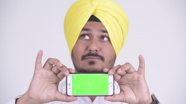 Face of happy Indian Sikh businessman thinking while showing phone - Footage, Video
