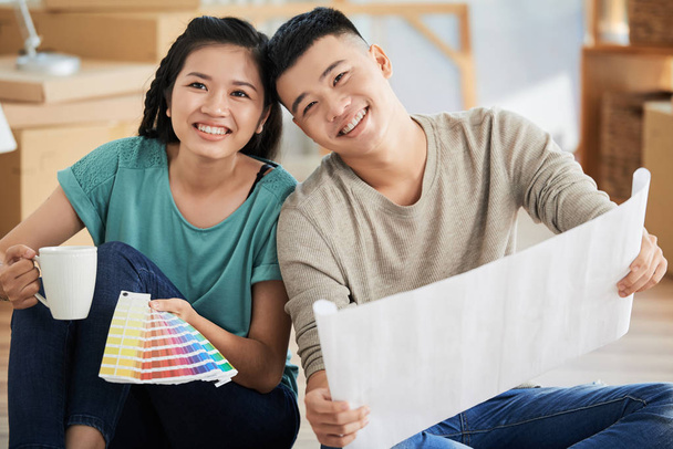 Mooie vrolijke Aziatische man en vrouw zitten op verdieping van nieuw appartement bedrijf cup en kleuren palet - Foto, afbeelding