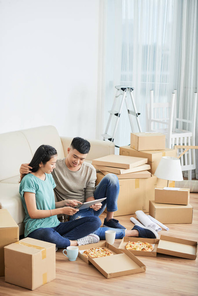 Alegre joven asiático hombre y mujer sentado en el piso de nuevo apartamento rodeado de cajas de cartón lleno y deliciosa pizza apoyada en el sofá y mirando a la tableta
  - Foto, Imagen