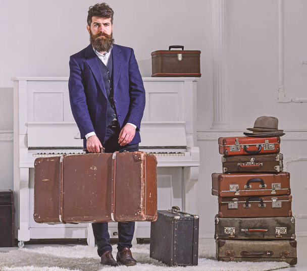 Macho elegant on strict face stands near pile of vintage suitcase, holds suitcase. Man, traveller with beard and mustache with luggage, luxury white interior background. Baggage delivery concept - Foto, Imagen