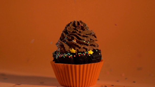 bunte Zuckerstreusel fallen auf dekorierten Cupcake mit Schokoladencreme auf orangefarbenem Hintergrund  - Filmmaterial, Video