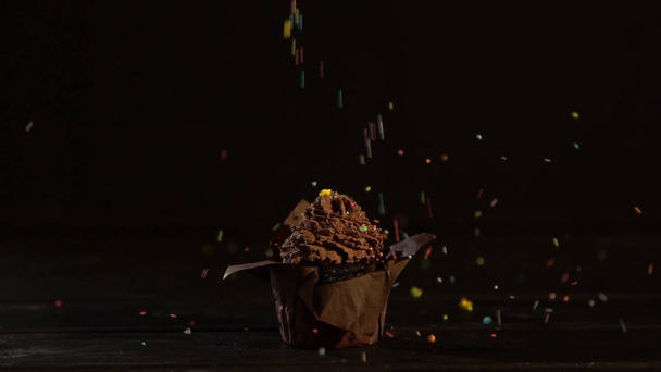 multicolored sprinkles falling down on cupcake in with chocolate cream on black background  - Filmagem, Vídeo