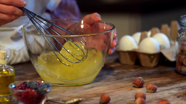 zwolnionym tempie kobieta kucharz ubijania jaj z trzepaczka w szklanej misce w drewniany stół - Materiał filmowy, wideo