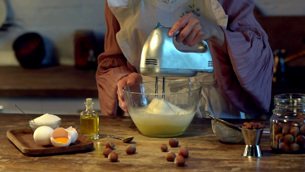 kobieta kucharz ubijania jaj w szklanej misce mikserem w drewniany stół z surowych składników - Materiał filmowy, wideo