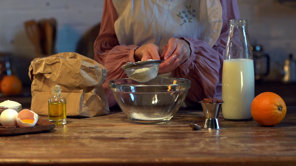 slow motion of female cook sifting flour in glass bowl at wooden table with ingredients - Footage, Video