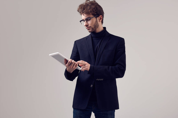 Fashion portrait of handsome elegant man with curly hair wearing suit and glasses holding a tablet on gray background in studio - Foto, immagini