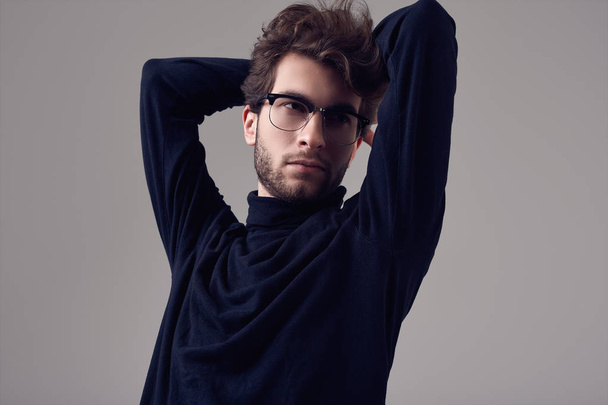 Fashion portrait of handsome elegant man with curly hair wearing black turtleneck and glasses on gray background in studio - Φωτογραφία, εικόνα