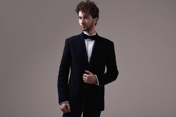 スタジオの灰色の背景にポーズのタクシードを着て巻き毛を持つハンサムなエレガントな男のファッション ポートレート - 写真・画像