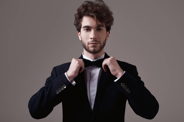 Fashion portrait of handsome elegant man with curly hair wearing tuxedo posing on gray background in studio - Fotoğraf, Görsel