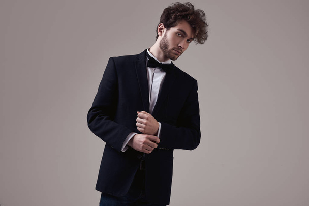 Fashion portrait of handsome elegant man with curly hair wearing tuxedo posing on gray background in studio - Photo, Image