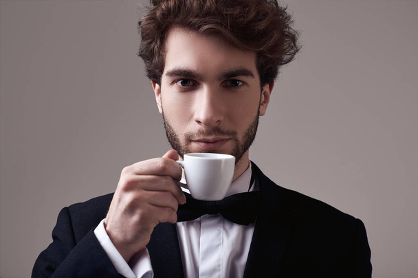 Fashion portrait of handsome elegant man with curly hair wearing tuxedo holding a cup of espresso on gray background in studio - Photo, image
