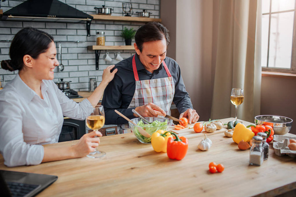 Man sit at table in kitchen and cut vegetables for salad. He look down and smile. His wife support him by holding hand on shoulder. She smile. Woman has glass of wine in hand. - Fotó, kép