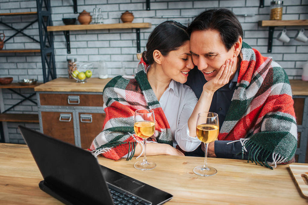Womn sit with man at table in kitchen. She hold her hand on his cheek and lean to him. Man look on laptop. They have glasses of wine on table. - 写真・画像