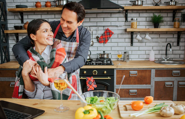 Nice couple is in kitchen. Guy stand behind woman and embrace her. She has blanket on shoulders. Woman sit at table and hold husband hands in her. - Photo, image