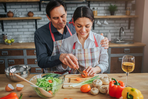 Nice man stand behind woman in kitchen. He help her to cut tometoes. Woman look down and smile. They hold vegetables and glasses of wine on table. - Photo, image