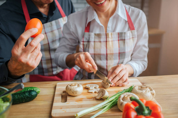 Geknipte weergave van mans hand met tomaat. Vrouw zitten aan tafel en gesneden champignons. Smhe glimlacht. Ze zijn in de keuken. - Foto, afbeelding