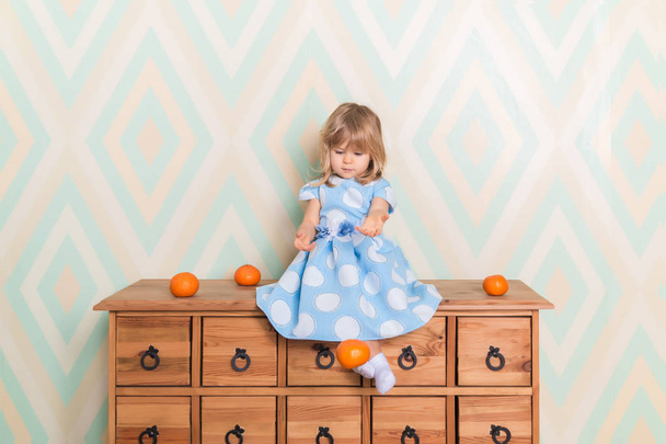 Little baby girl in her room sitting cross-legged on chest of drawers and throws tangerines down on rhomb wallpaper background. Child in blue polka dot dress and white socks attentively looking down - Foto, Bild