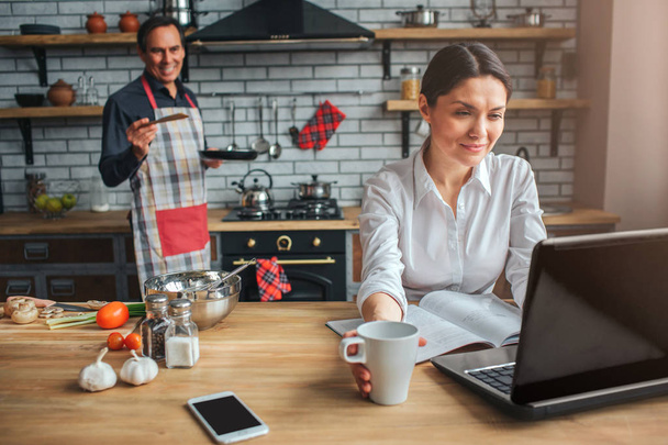 Woman sit at table in kitchen. She look at laptop and work. Woman smile. Man stand at stove. He hold frying pan and look at woman. Guy smile. - Photo, image