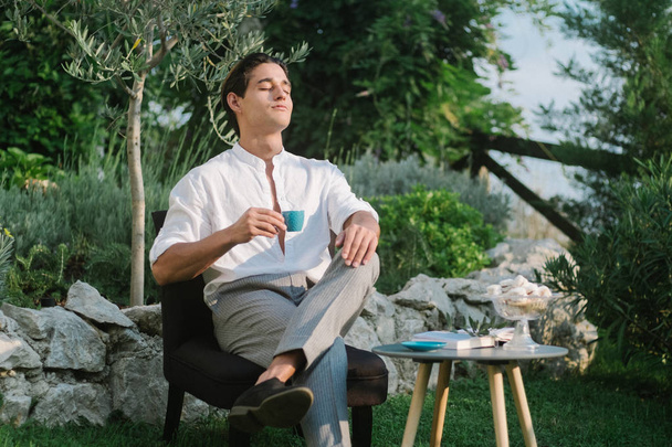 Attractive young fashion man in a suit drinks coffee in the morning and reads a book outdoors in the garden.  - Foto, Bild