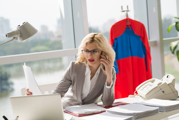 Atraktivní podnikatelka kavkazských rozhovor na smartphone a zároveň sedí v kanceláři s papíry a laptop, kostým superwoman visí za - Fotografie, Obrázek