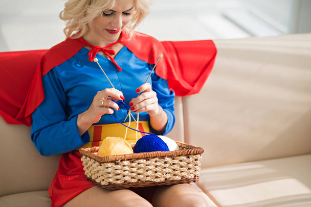 Young beautiful blond Caucasian woman wearing superwoman costume knitting while sitting on couch with basket of yarn - Foto, imagen