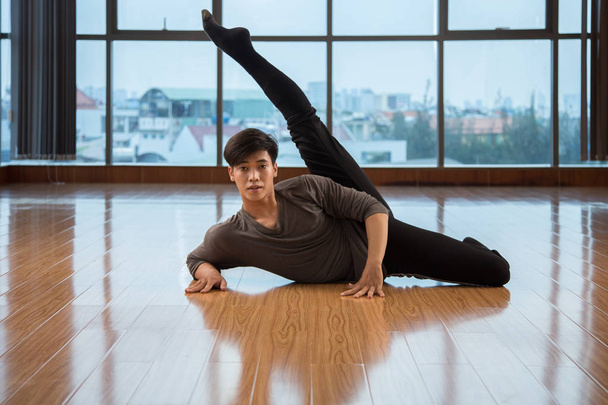Atraente asiático masculino levantando perna e olhando para a câmera enquanto dança no chão no estúdio moderno
 - Foto, Imagem
