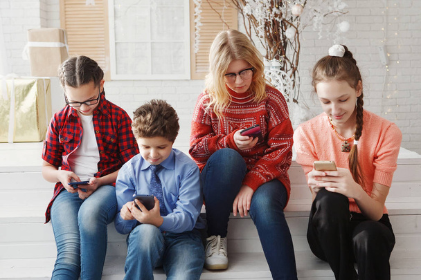 Czterech przyjaciół z wesoły Nastolatka siedzi na białych drewnianych schodkach, uśmiechając się, patrząc na smartfony, rozmowy w sieciach społecznościowych. Przyjaźń, młodość, radość, komunikacja, Internet. - Zdjęcie, obraz