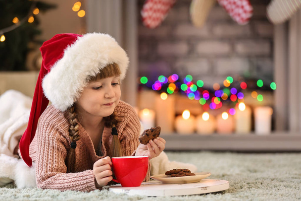 Милая маленькая девочка в шляпе Санты пьет горячий шоколад и ест печенье дома в канун Рождества
 - Фото, изображение