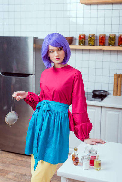 schöne Hausfrau mit lila Haaren und bunten Kleidern, die Sieb hält und in der Küche posiert - Foto, Bild