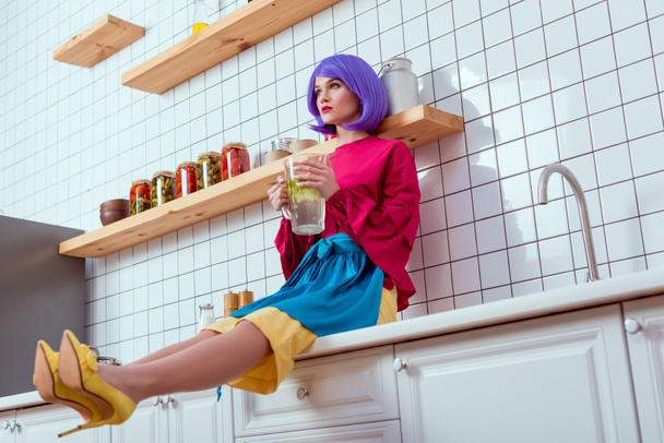 selective focus of housewife with purple hair and colorful clothes sitting on kitchen counter and holding glass jar of lemonade - Foto, afbeelding