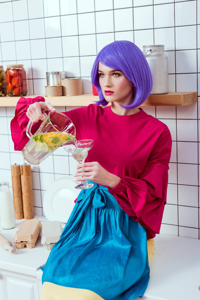 housewife with purple hair and colorful clothes sitting on kitchen counter and pouring lemonade in glass - Photo, Image